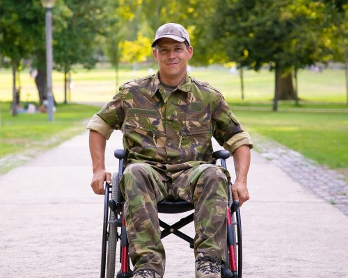Happy disabled military man in wheelchair wearing camouflage uniform, moving on footpath in city park. Front view. Veteran of war or disability concept
